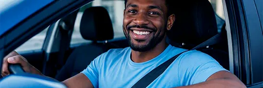 Homem negro, vestindo uma camiseta azul, sentado no banco do motorista do carro, com o braço apoiado na janela e sorrindo.