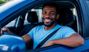 Homem negro, vestindo uma camiseta azul, sentado no banco do motorista do carro, com o braço apoiado na janela e sorrindo.