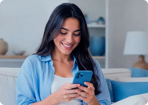 Mulher branca, vestindo uma camisa azul, sentada em um sofá com o celular nas mãos e sorrindo.