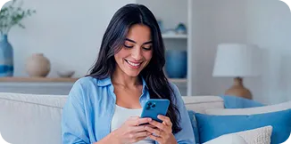 Mulher branca, vestindo uma camisa azul, sentada em um sofá com o celular nas mãos e sorrindo.
