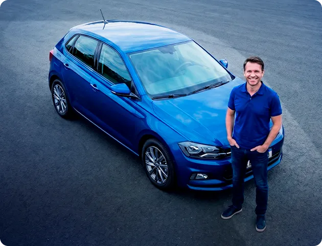 Homem branco em pé, com as mãos no bolso e vestindo uma camisa azul. Logo atrás dele está um carro azul.