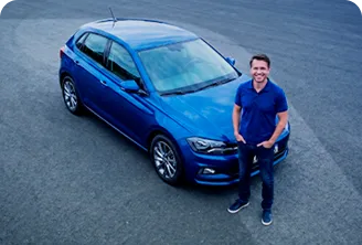 Homem branco em pé, com as mãos no bolso e vestindo uma camisa azul. Logo atrás dele está um carro azul.