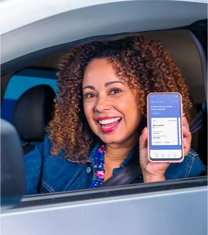 Mulher sentada na posição do motorista, sorrindo na janela com o celular aberto no aplicativo do Banco BV