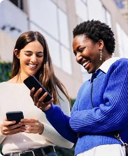 Duas mulheres sorrindo ao ar livre olhando celulares em frente a prédio.