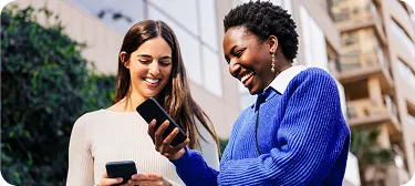 Duas mulheres sorrindo ao ar livre olhando celulares em frente a prédio.
