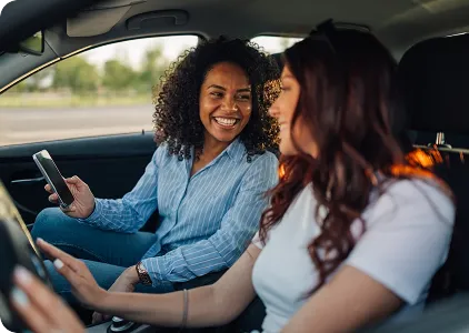 Duas mulheres dentro de um carro, uma sorrindo e segurando um celular enquanto a outra dirige.