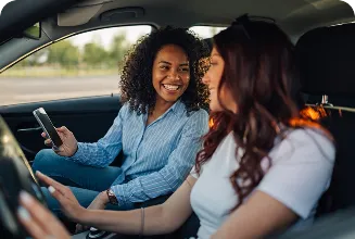 Duas mulheres dentro de um carro, uma sorrindo e segurando um celular enquanto a outra dirige.
