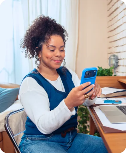 Uma mulher está sorrindo enquanto olha o celular e o segura com uma das mãos.