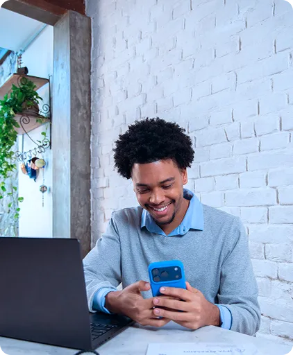 Um homem sorri enquanto segura e olha para o celular, com as duas mãos apoiadas sobre a mesa, na sua frente um notebook.