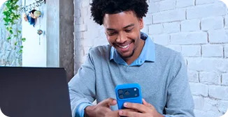 Um homem sorri enquanto segura e olha para o celular, com as duas mãos apoiadas sobre a mesa, na sua frente um notebook.