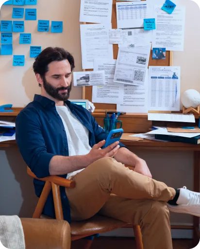 Um homem está com a cabeça abaixada, sentado em uma cadeira, olhando o seu celular. Uma das pernas está cruzada.