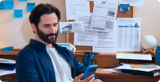 Um homem está com a cabeça abaixada, sentado em uma cadeira, olhando o seu celular. Uma das pernas está cruzada.