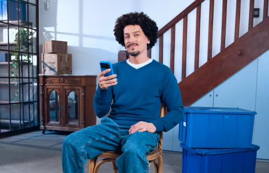 Um homem sorri para a câmera enquanto está sentado em uma cadeira e segura o celular com uma das mãos.