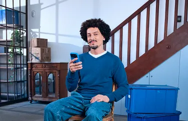 Um homem sorri para a câmera enquanto está sentado em uma cadeira e segura o celular com uma das mãos.