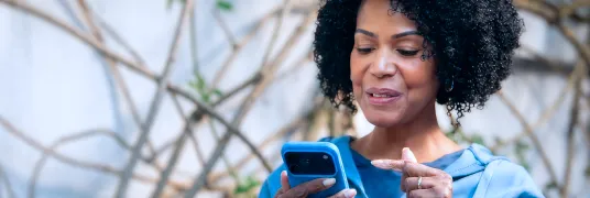 Em um plano fechado, uma mulher sorri enquanto olha para o celular e aponta para a tela com uma das mãos.