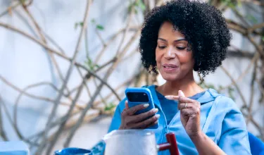 Em um plano fechado, uma mulher sorri enquanto olha para o celular e aponta para a tela com uma das mãos.
