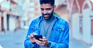 Um homem de jaqueta azul, segura o celular com as duas mãos, olha para a tela e sorri.