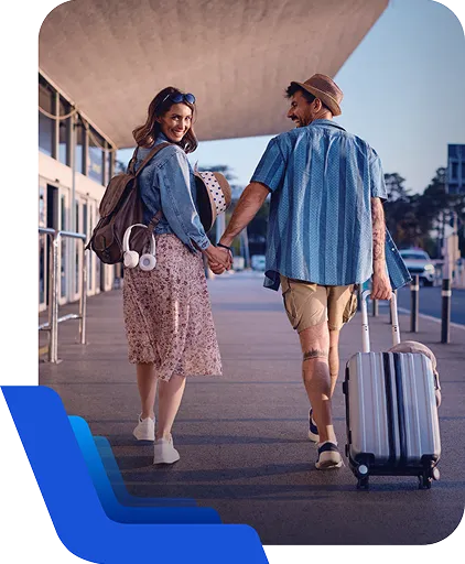 Duas pessoas caminham de mãos dadas com malas em área externa, em contexto de viagem.