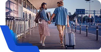 Duas pessoas caminham de mãos dadas com malas em área externa, em contexto de viagem.