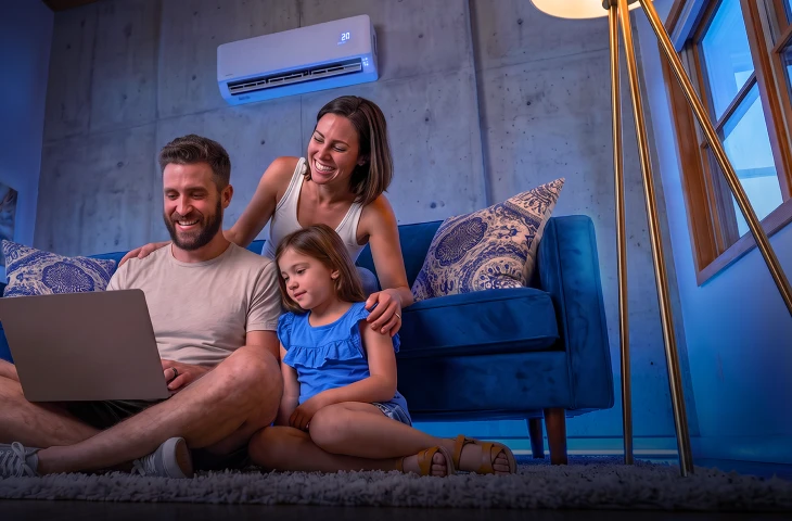Mulher em sofá azul apoia as mãos nos ombros de um homem e de uma menina, enquanto acompanham juntos algo no computador.