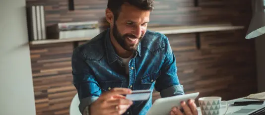 Homem de cabelo curto e barba, usando uma camisa de botão azul. Ele segura com uma mão um cartão BV e com a outra um tablet. Acesse o site do banco BV e fique por dentro das novidades!