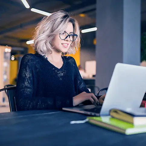 Mulher loira, com óculos e blusa pretos. Ela está mexendo atentamente em um notebook branco.