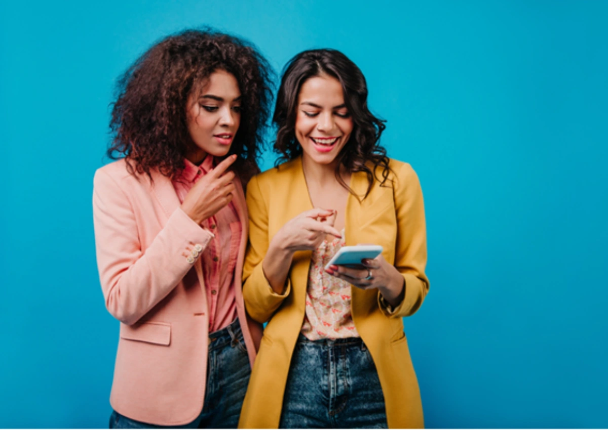 Mulheres verificando em smartphone como quitar dívidas no Feirão Negocia Já do BV, em ambiente com fundo azul. 