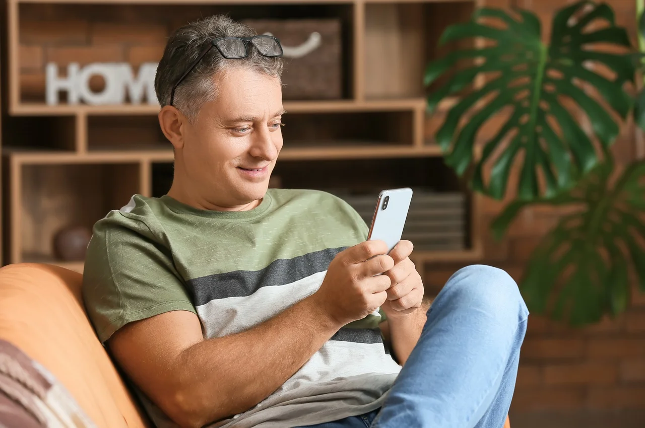 Homem sentado no sofá, utilizando o celular, com fundo de decoração de casa. 