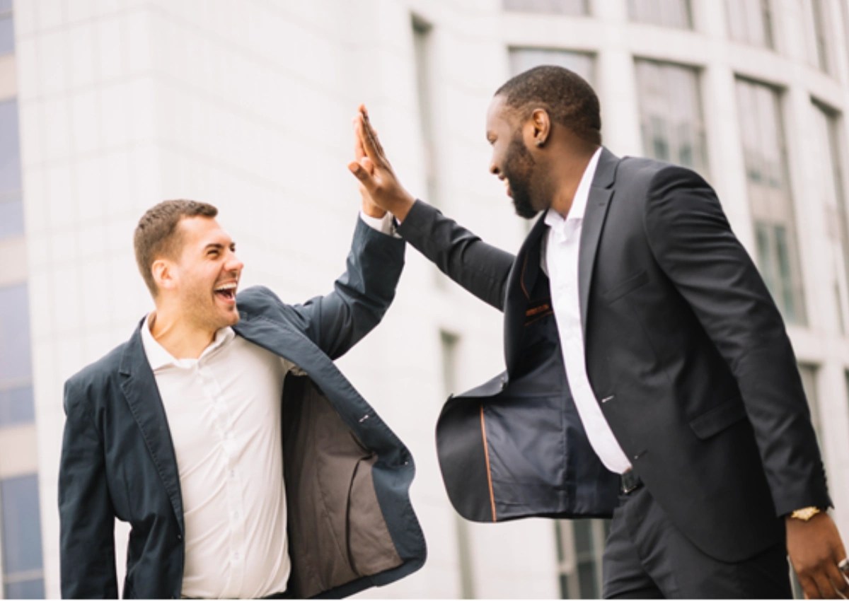 Homens felizes fazendo um high-five após quitar dívidas no Feirão Negocia Já do BV, em ambiente aberto, com um prédio ao fundo.