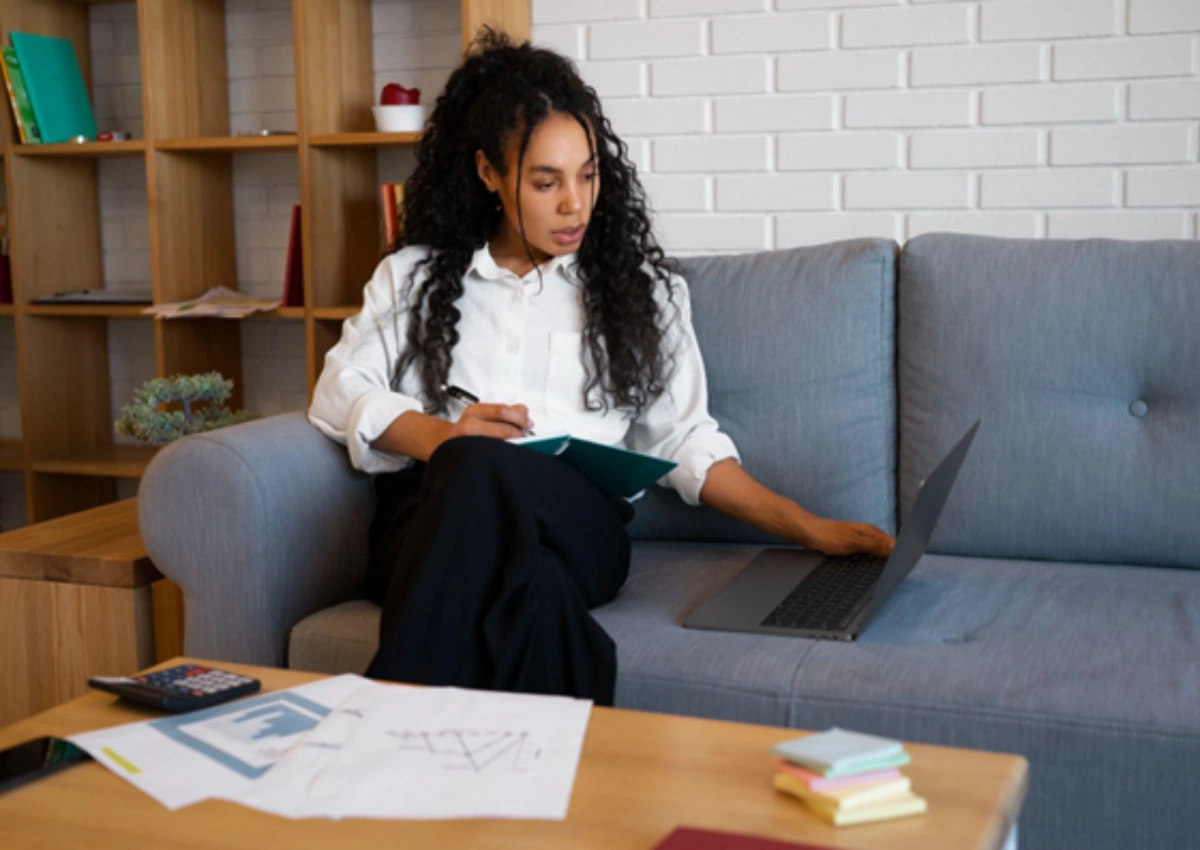 Mulher sentada em um sofá olhando para um notebook ao seu lado, com uma caneta na mão e prancheta no colo.