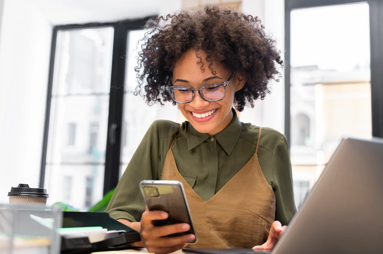 Mulher sorrindo com celular na mão, sentada em um ambiente de escritório. 