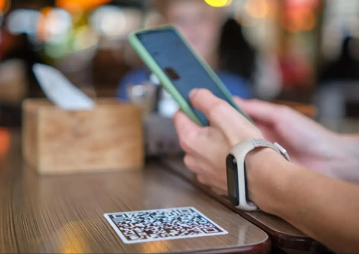 Duas mãos, apoiadas em uma mesa de madeira com QR Code em sua borda, segurando um celular.