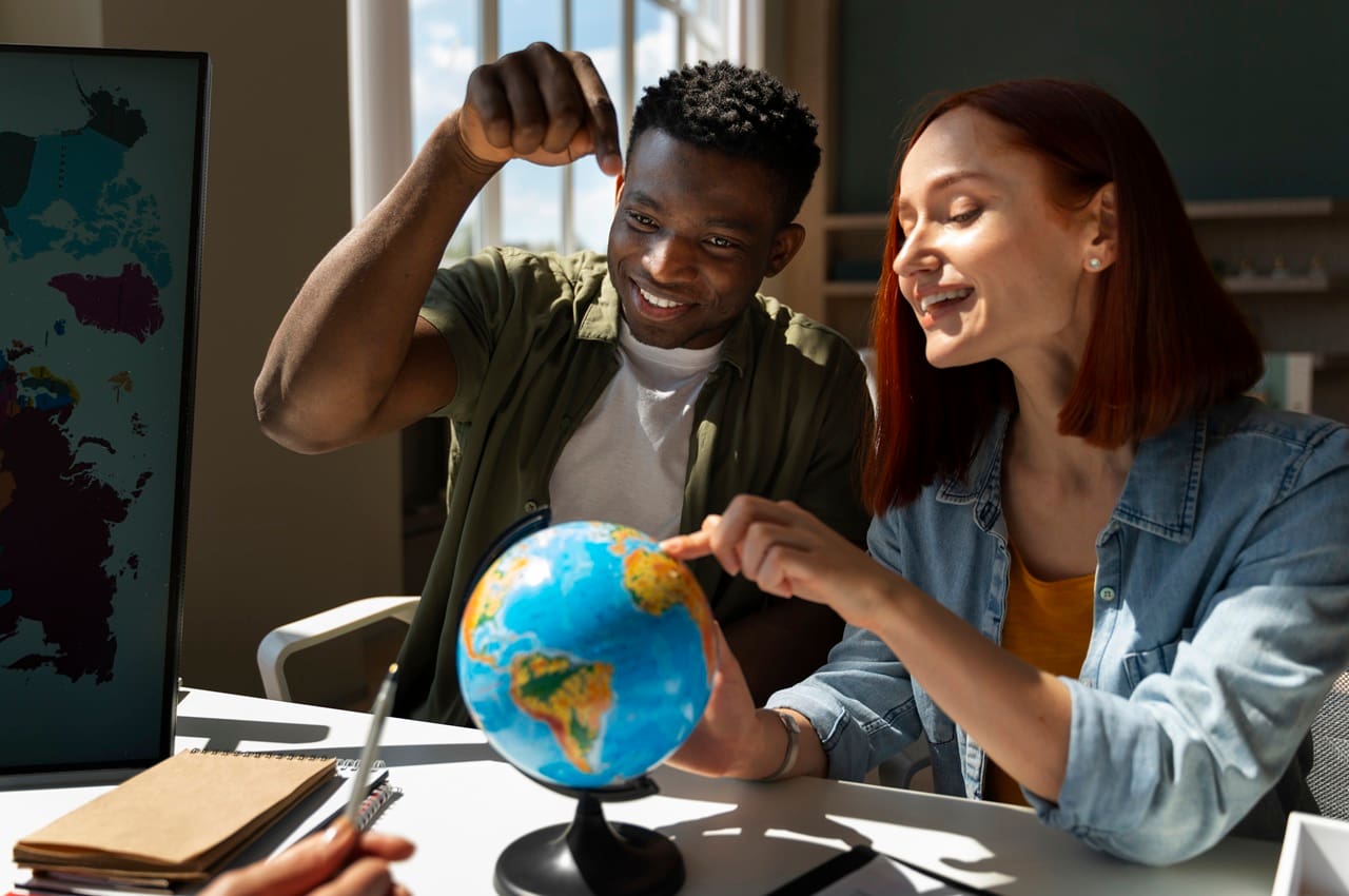 Dois jovens em uma mesa de escritório, observando um globo terrestre. Eles estão planejando um intercâmbio.