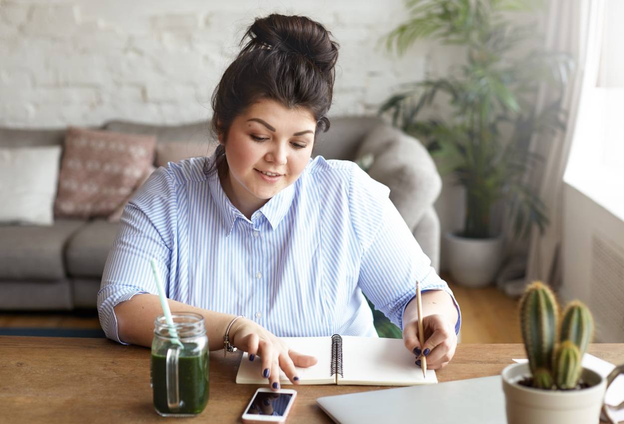 Mulher sentada à mesa anotando em caderno enquanto consulta o celular, ilustrando como usar o Open Finance para organizar as contas e melhorar a rotina financeira.