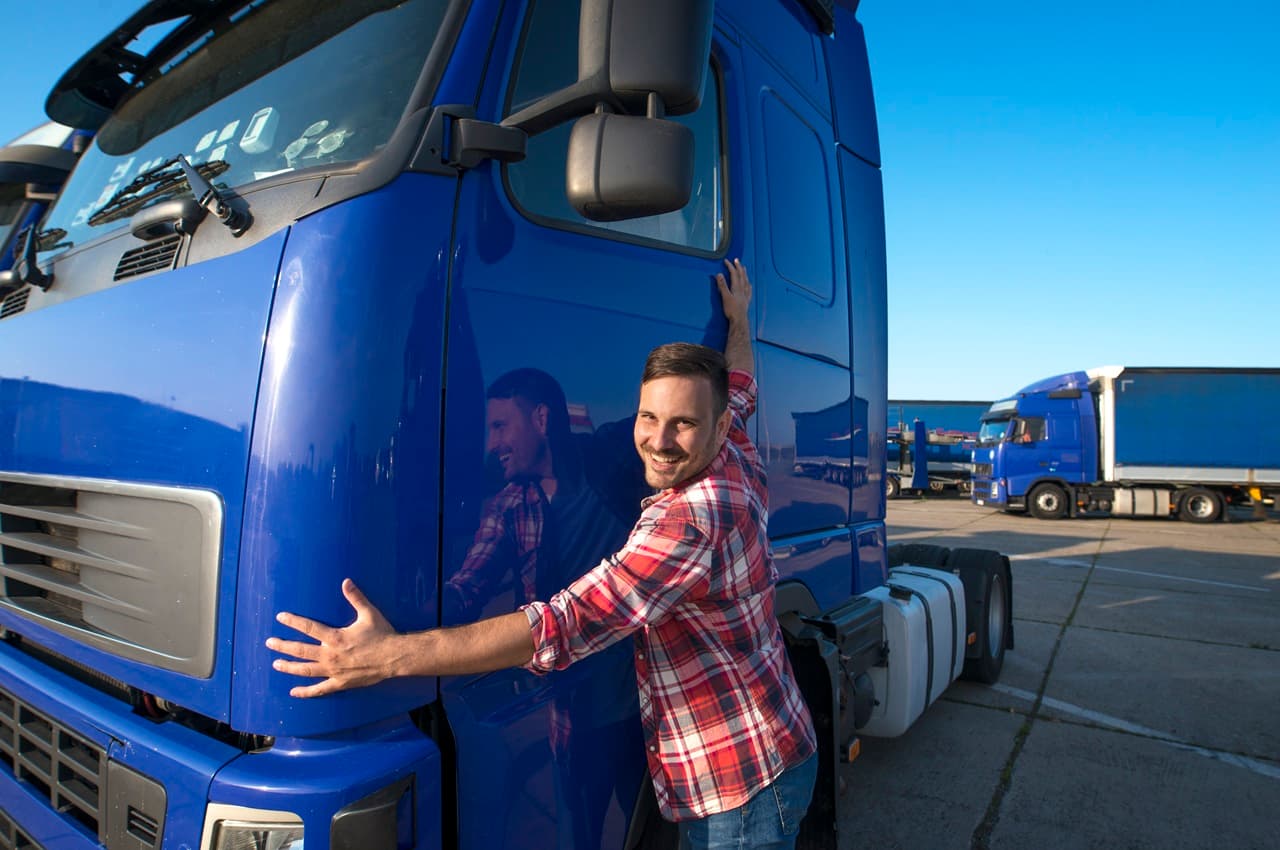Homem sorridente abraçando a frente de um caminhão azul em um pátio de transporte. Ele aproveitou os benefícios parar financiar um caminhão.