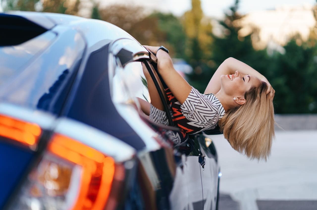 Mulher loira sorridente inclinando o corpo para fora da janela dianteira de um carro azul, com a cabeça para trás e a mão no cabelo.