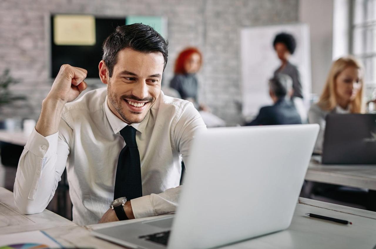 Homem sorridente em um ambiente de escritório. Ele está olhando para a tela de um notebook e fazendo um sinal afirmativo com uma das mãos.