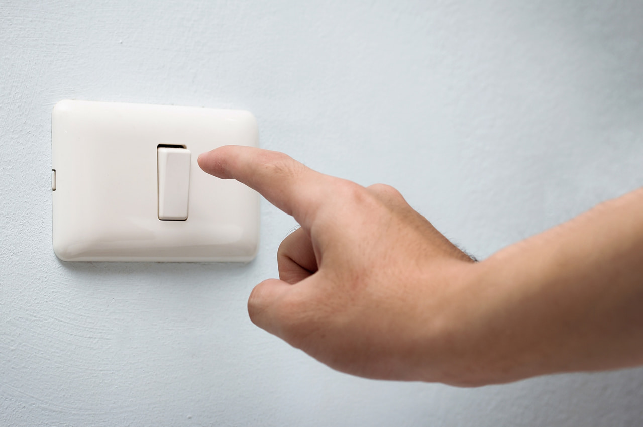 Pessoa prestes a desligar o interruptor de luz em uma parede branca, sinalizando como evitar desperdício de energia.