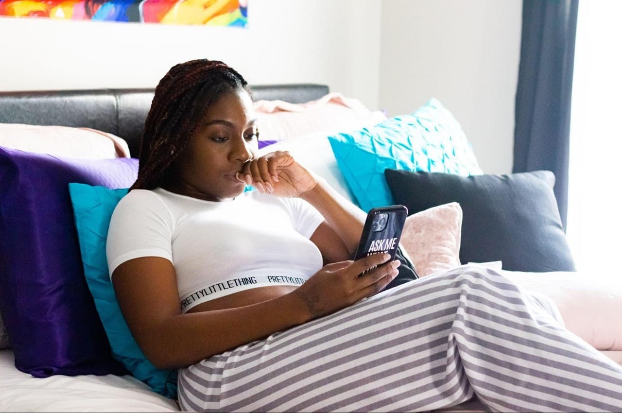 Mulher negra com expressão concentrada, apoiada em almofadas coloridas azuis na sua cama e utilizando celular com apps de controle financeiro. A mulher veste cropped branco e calça listrada cinza. 