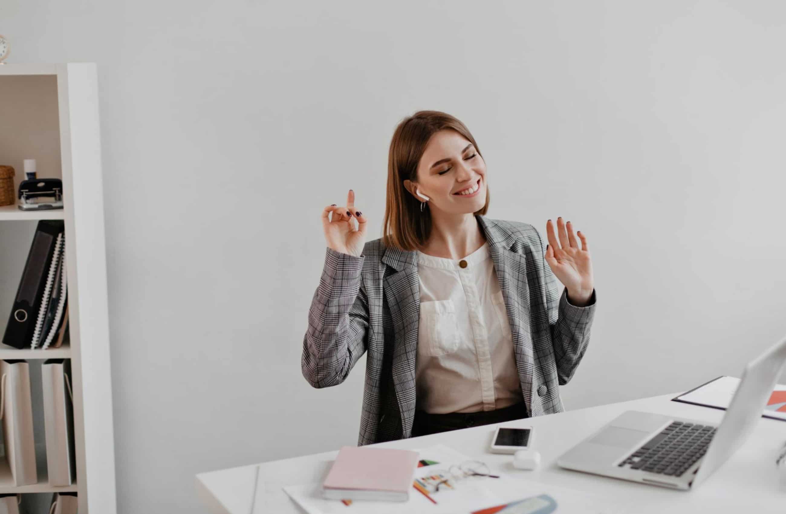 Uma mulher jovem e sorridente, usando blazer cinza e fones de ouvido sem fio, gesticula levemente enquanto trabalha em um escritório iluminado. A imagem transmite a tranquilidade de ter uma vida financeira organizada.