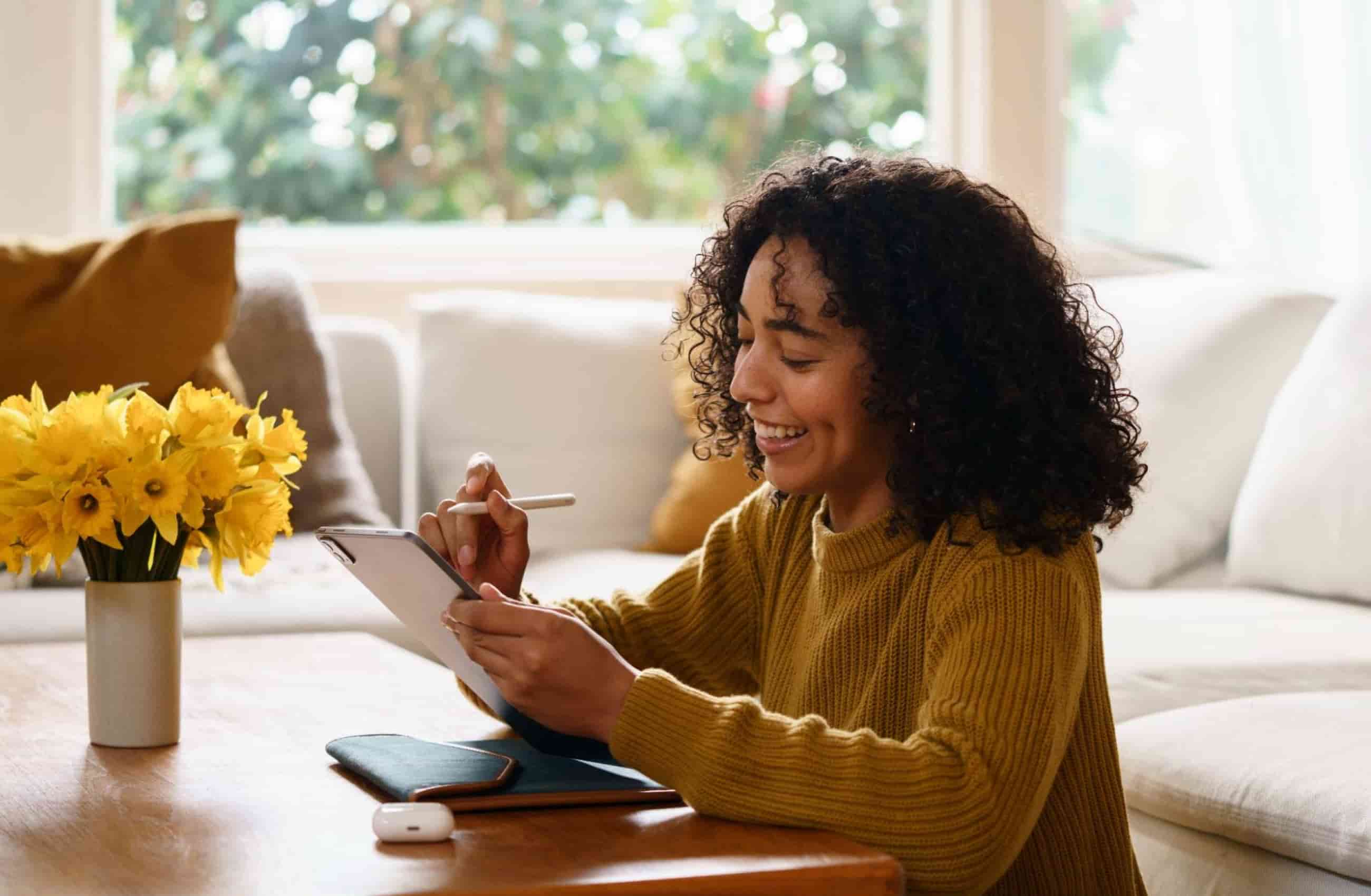 Uma mulher sorridente com cabelos cacheados utiliza um tablet com uma caneta digital em uma sala iluminada. A cena ilustra a facilidade de organizar finanças e gerenciar contas com a Assistente de Pagamentos BV.