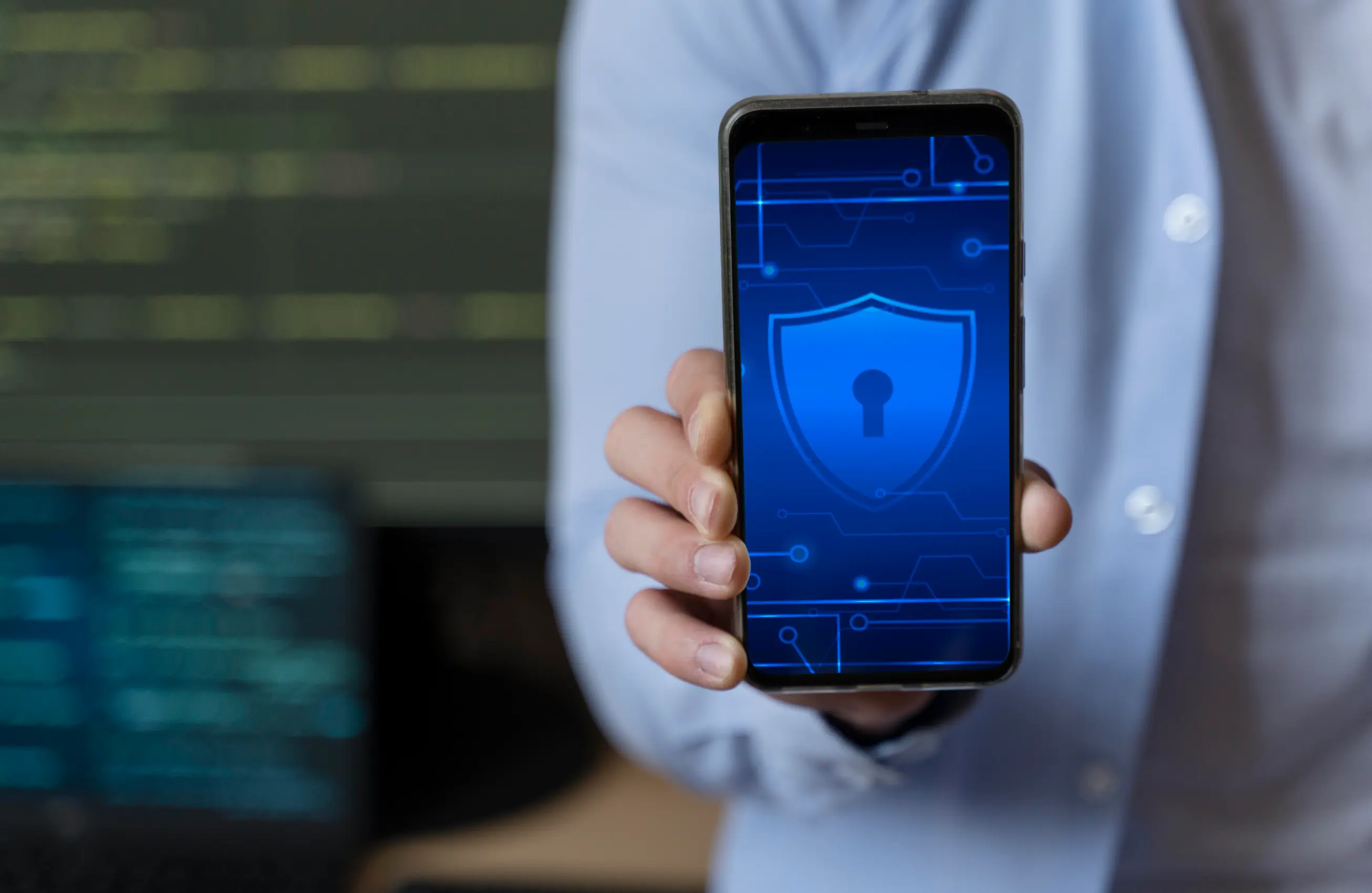 Pessoa mostrando um smartphone com a tela voltada para a câmera exibindo um ícone de escudo azul com cadeado, sugerindo segurança digital; fundo desfocado com tela de computador. 