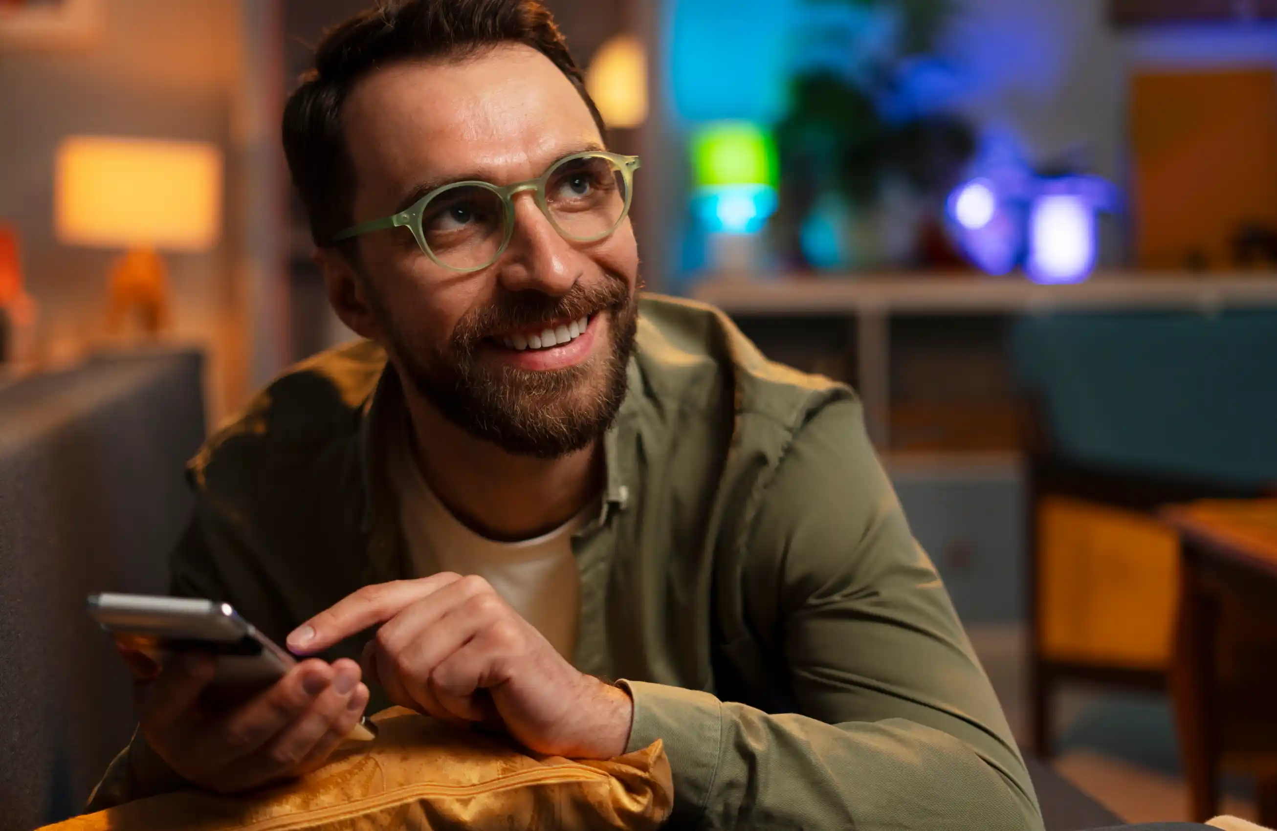 Um homem utilizando um smartphone enquanto sorri para algo fora da imagem. Ele aparenta estar em uma cafeteria ou sala de estar.
