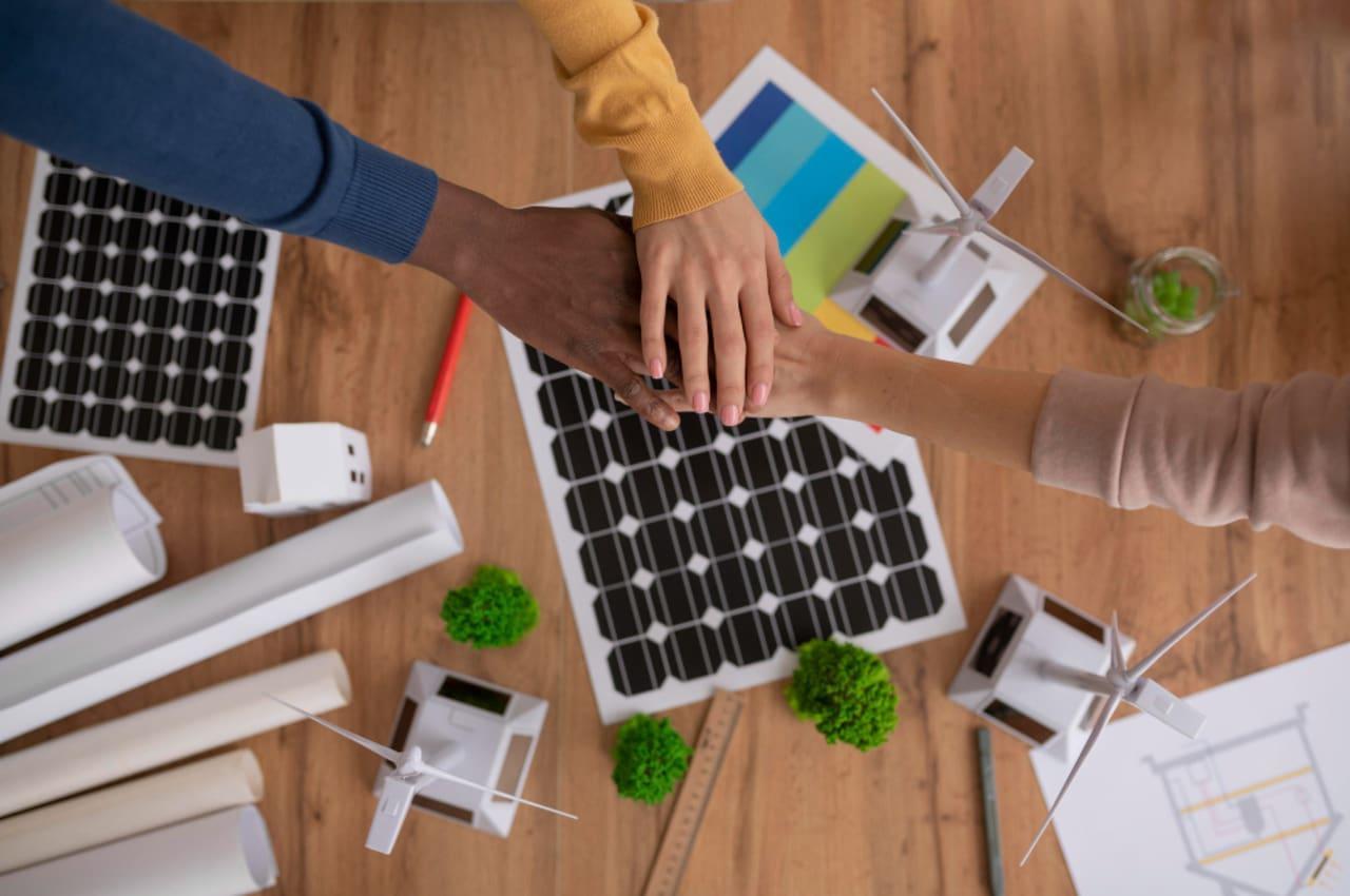 Mãos se unindo sobre uma mesa com materiais de energia solar, incluindo painéis solares e mini turbinas eólicas.