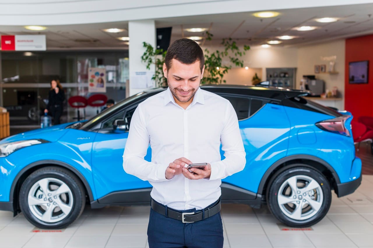 Homem sorridente segurando um celular, com gesto de celebração de uma conquista. Ele está em frente a um carro azul.