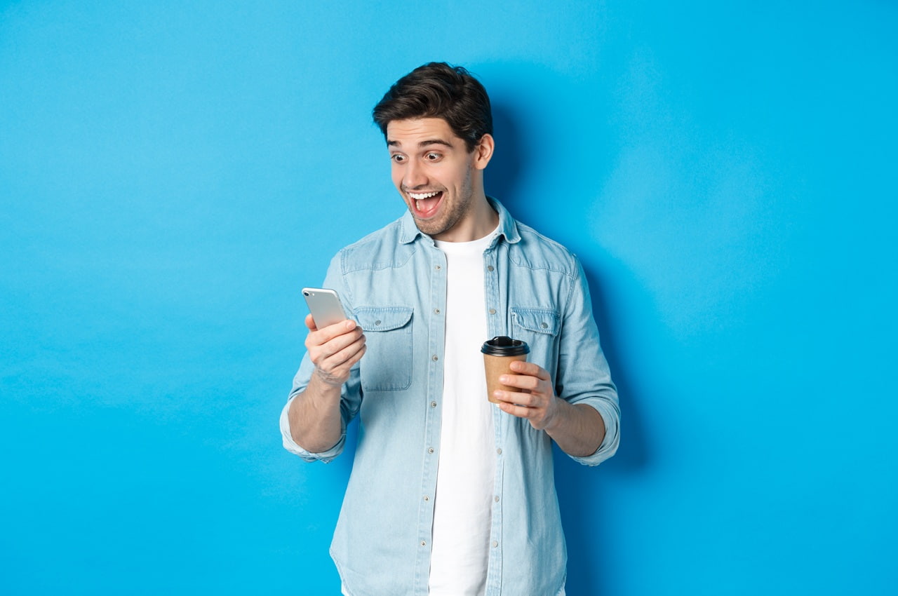 Homem jovem sorridente em frente a um fundo azul. Ele está com um copo de café em uma das mãos e com um celular na outra, contratando um projeto de energia solar pelo Feirão Solar BV.