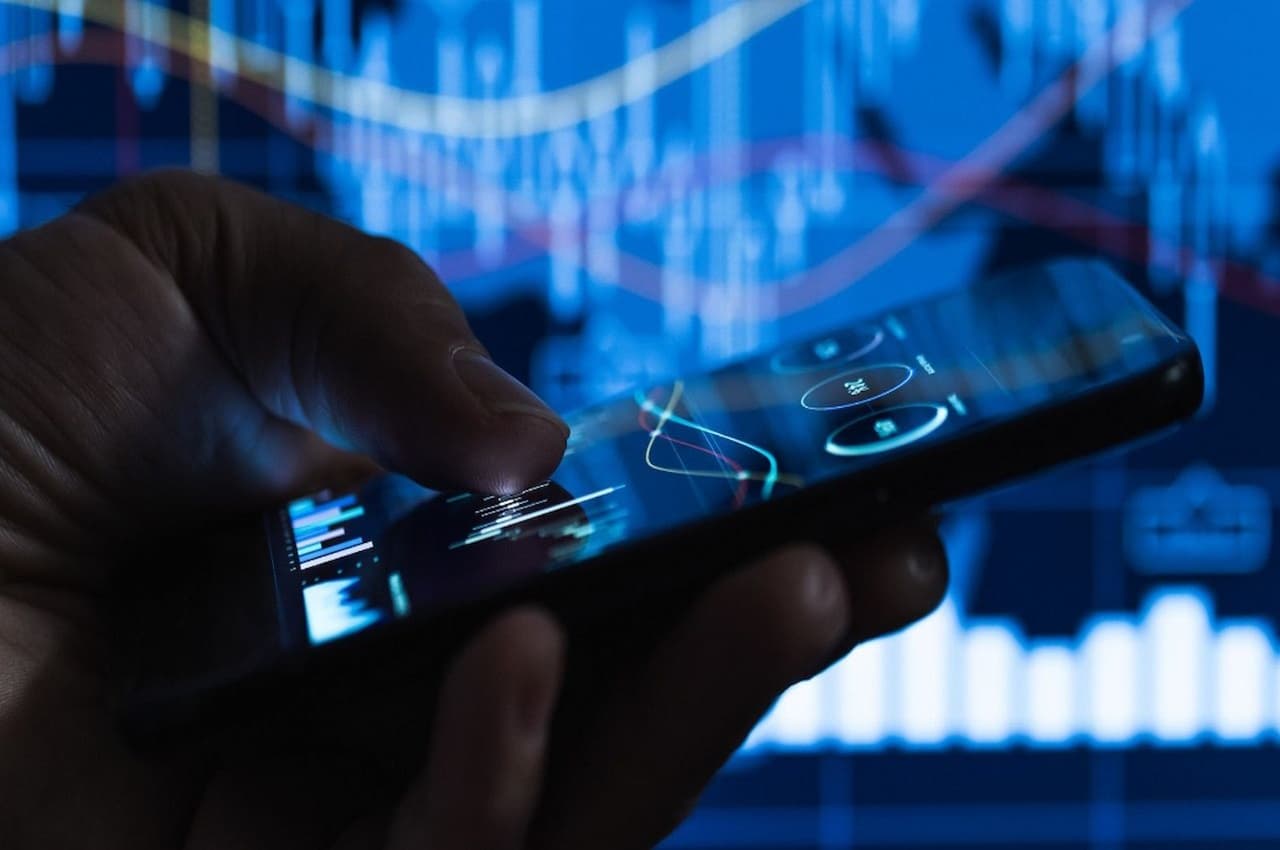 Foco em mão acessando blockchain pelo celular. A tela do aparelho mostra valores (não é possível ver) e linhas suaves em um gráfico.