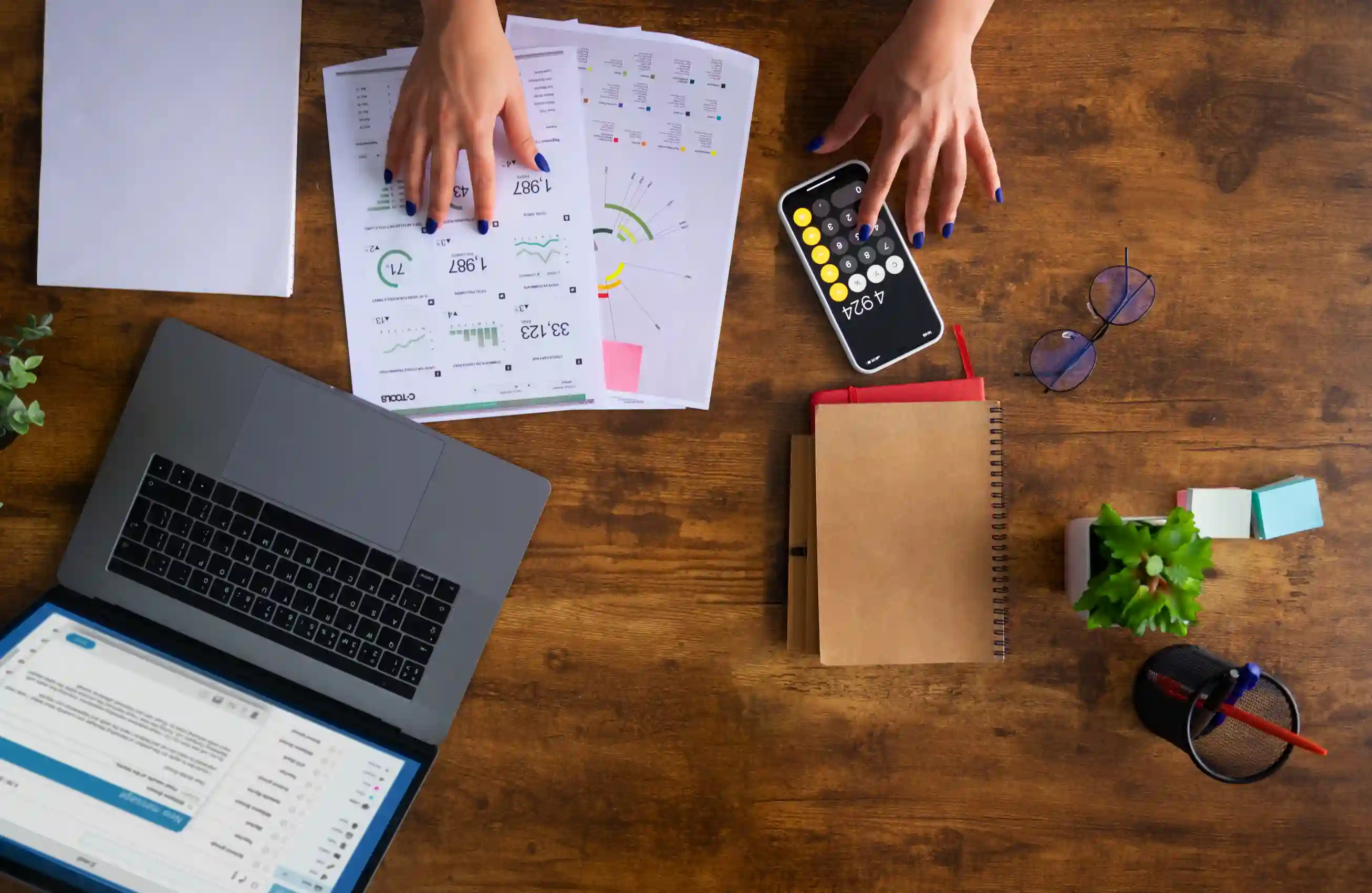 Uma imagem de uma mesa de escritório capturada de cima. Nela, estão espalhados um notebook, caderno, folhas com dados e um smartphone com calculadora.