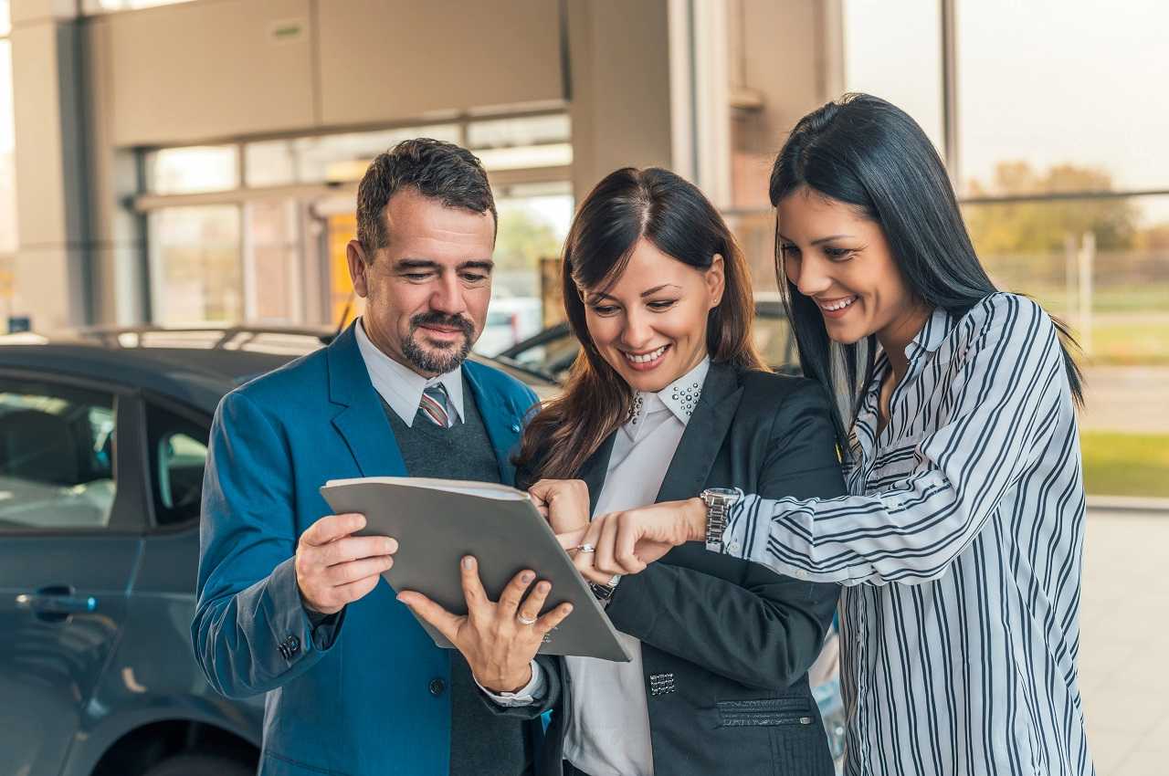 Grupo de três pessoas com um homem e duas mulheres sorridentes. O grupo está em pé em uma concessionária olhando para uma pasta cinza nas mãos de uma mulher que veste blazer cinza.