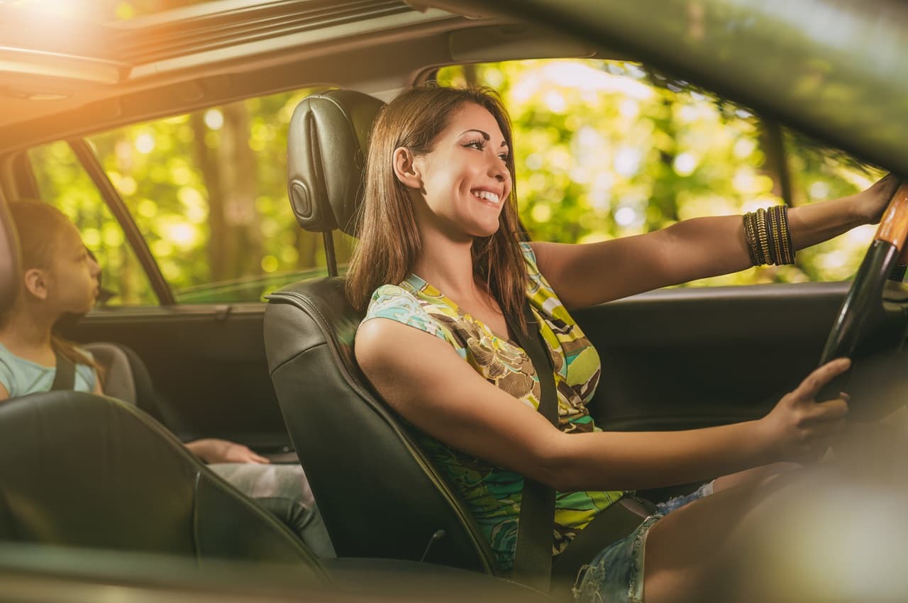 Mulher sorridente vestindo blusa florida e shorts jeans, dirigindo seu carro ideal e levando sua filha no banco passageiro.